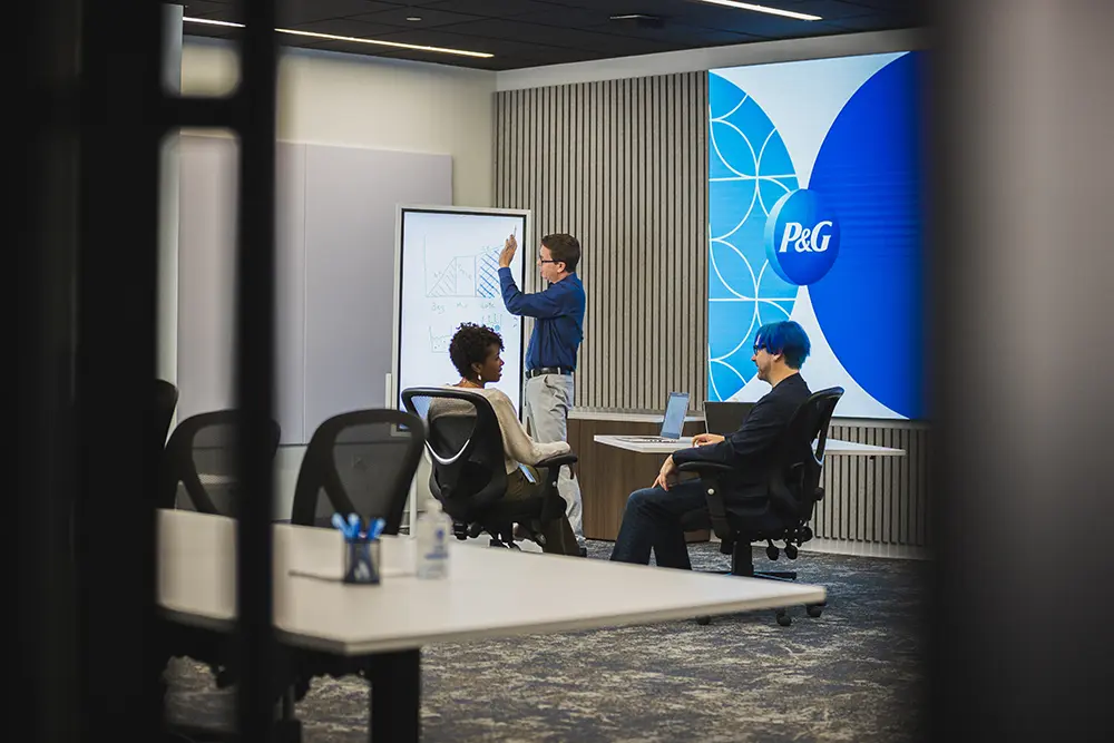 A man with light skin wearing glasses and a navy shirt draws graphs on a whiteboard. In the foreground, a woman with warm brown skin and curly hair, and a man with light skin, blue hair and glasses sit and chat.