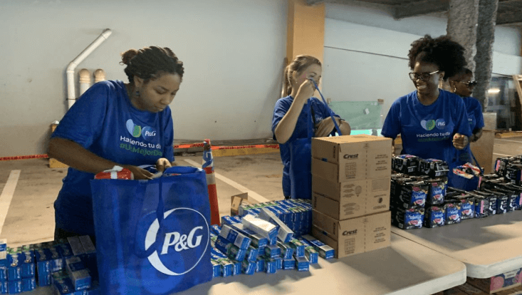 Three women wearing dark blue t-shirts are unboxing P&G products and packing large blue tote bags. They are standing behind a table full of products. 