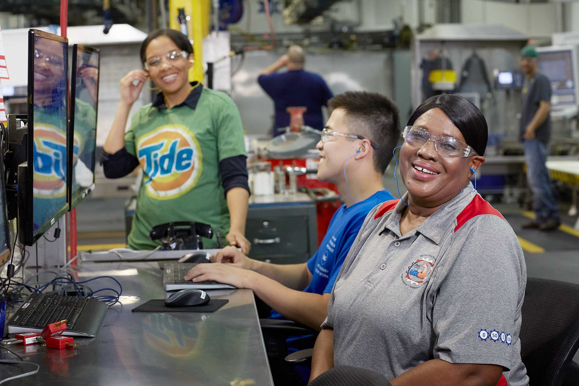 Two women and a man sit in front of several computers in a manufacturing plant. They wear protective eye gear and smile at the camera.