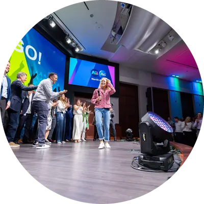 A small crowd of men and women clap and cheer on a blond-haired woman in jeans and a pink jacket as she walks across a stage.