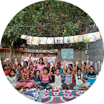 A large group of children sit on colorful mats outdoors with their hands raised, surrounded by hanging books, drawings, and chalkboard art against a garden backdrop.
