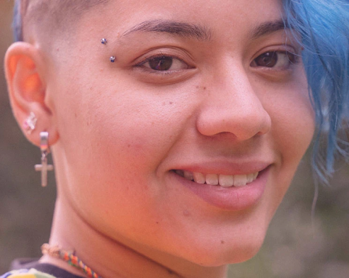 Girl smiling towards the camera. She has blue hair and part of her hair shaved.