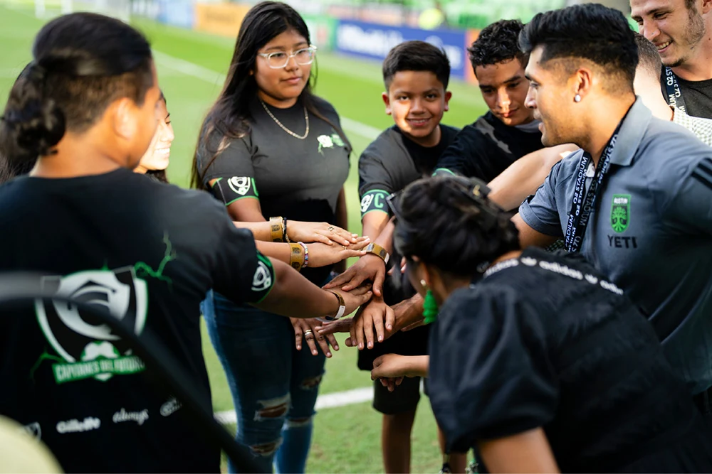 Seven Hispanic adolescents stand around in a circle. They are wearing black t-shirts and holding hands together in the center of their circle.