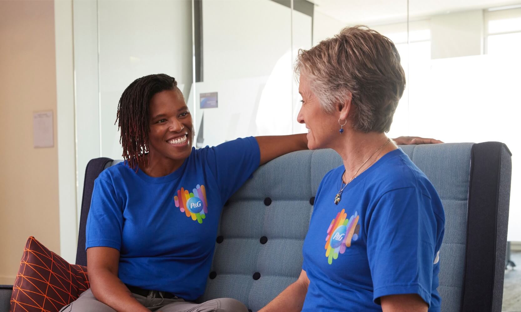 Two smiling women wearing t-shirts with a heart shaped Pride logo and the P&G logo sit together on a sofa.