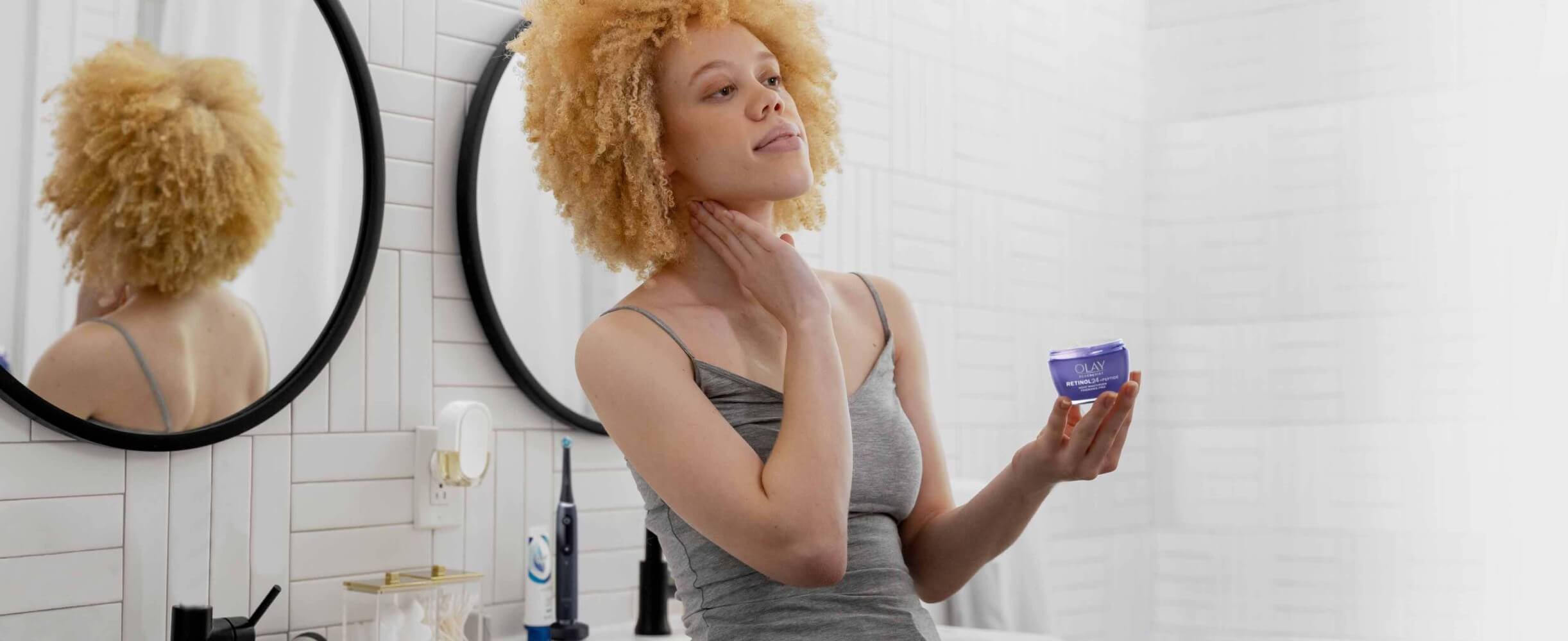 Woman in white tiled bathroom applies Olay Retinol 24 cream. Oral-B iO toothbrush and Crest toothpaste tube are on bathroom counter in background.  