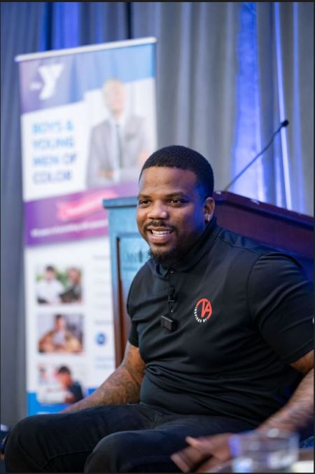 A Black man in black attire smiles while seated near a stage