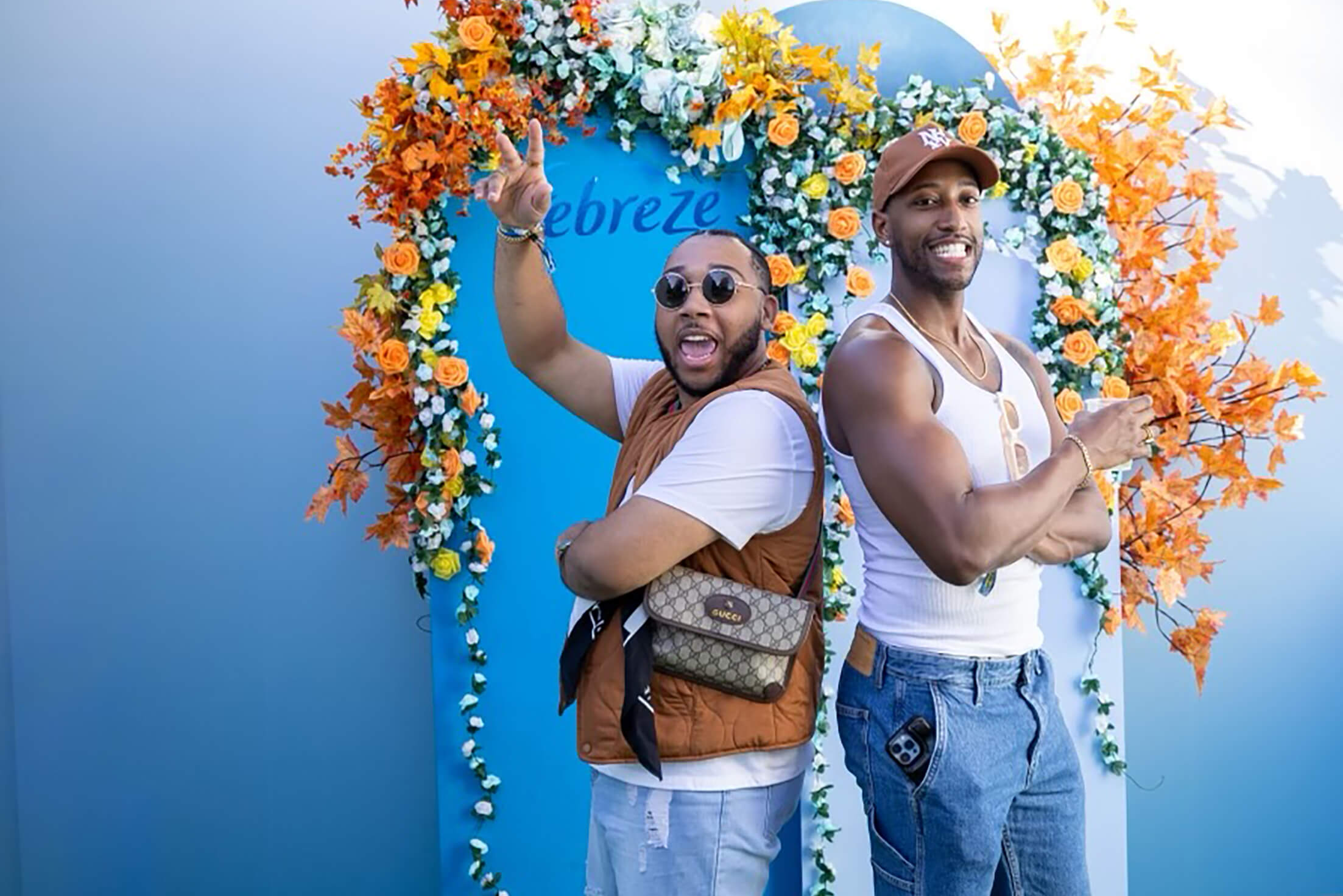 Two young Black men pose back-to-back. They stand in front of a sign decorated with flowers and text that says, "Febreze."