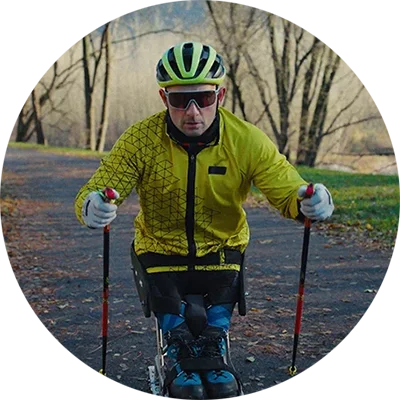 A man in a bright yellow jacket, green helmet, and sunglasses practices Para cross-country skiing on a paved path, holding ski poles and using adaptive equipment. Bare trees line the path, and mountains are visible in the distant background.