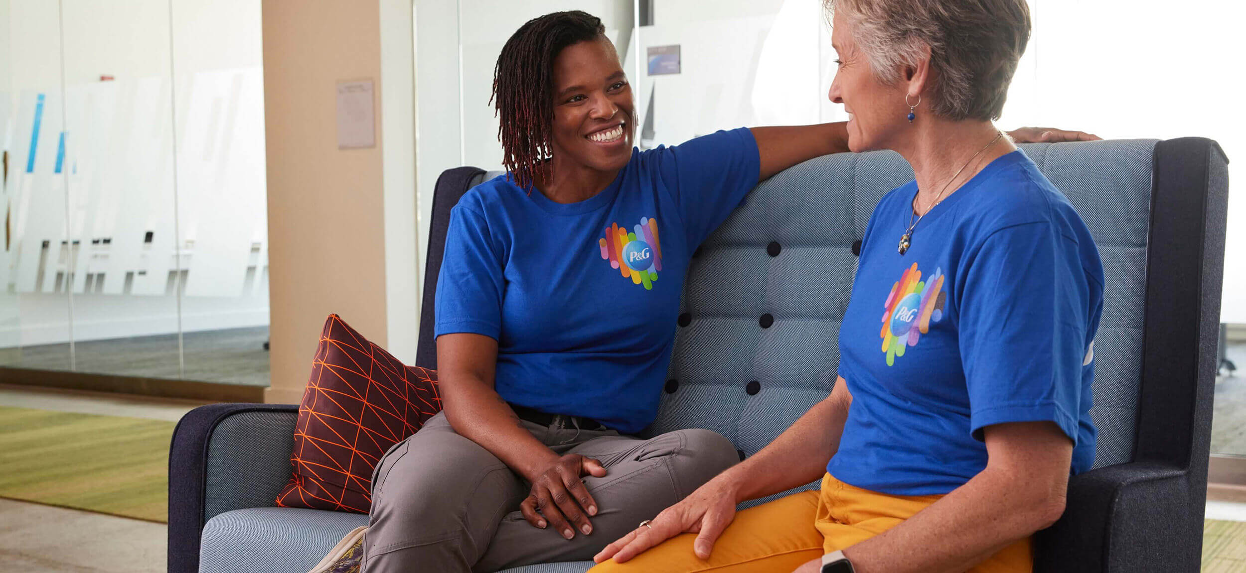 Two smiling women wearing t-shirts with a heart shaped Pride logo and the P&G logo sit together on a sofa.