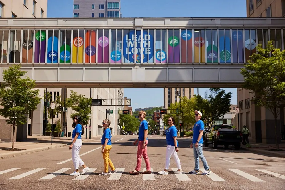 P&G employees cross street in front of rainbow Lead with Love sign featuring P&G brands.