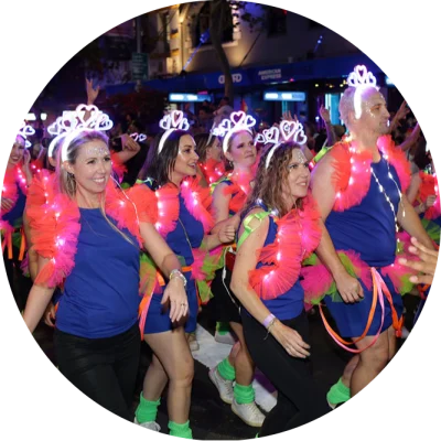 Over a dozen men and women smile as they march together in an evening parade. They are wearing matching outfits of blue shorts and shirts, pink feathered scarves and battery lit necklaces and crowns.