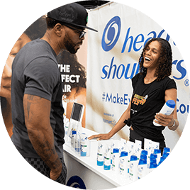 A young Black woman stands in front of a table lined with blue and white hair products. She holds one of the bottles as she engages with a young black man.