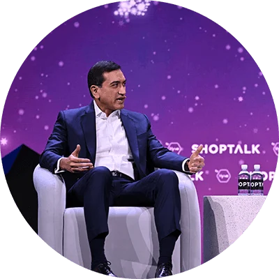 Two mean in dark blue business suits sit together on a stage while having a discussion. A purple digital background also displays a white text logo that reads, "Shoptalk."