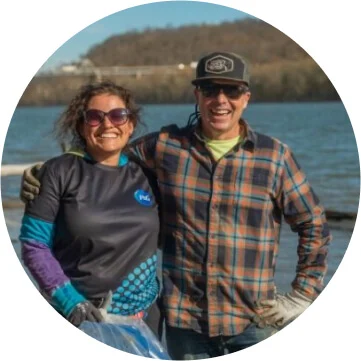 A man in a plaid shirt and baseball cap has his arm wrapped around a woman in a black, blue and purple shirt and sunglasses. They are both smiling towards the camera and there is a body of water behind them. 