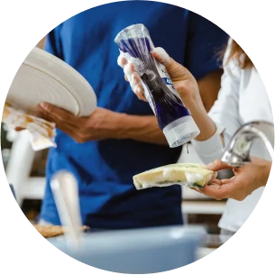 A person is squeezing dish soap onto a sponge and another person is drying a plate behind them.