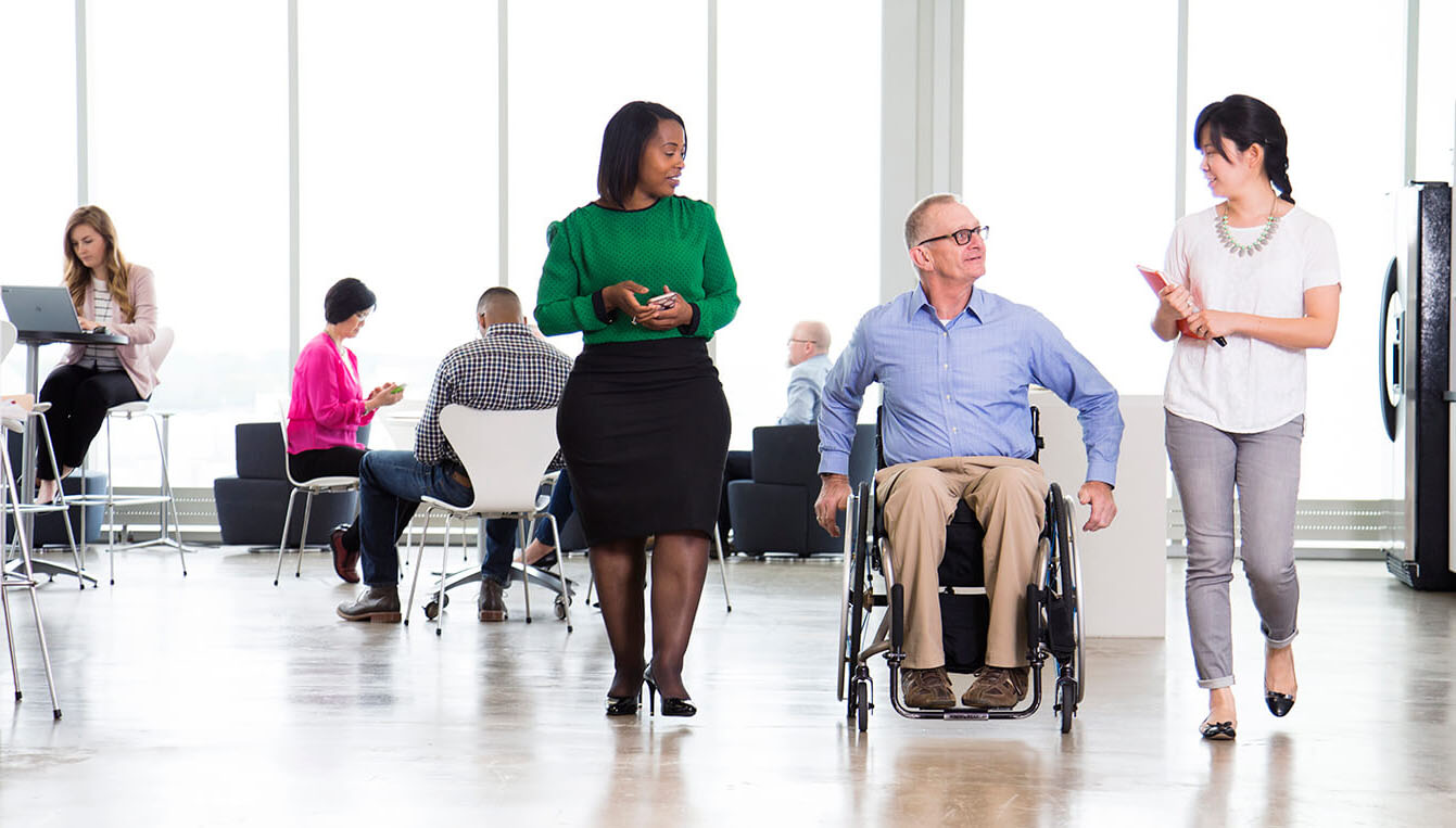 Two women walk alongside a man who is in a wheelchair. They are all dressed in business attire as they walk through an office.