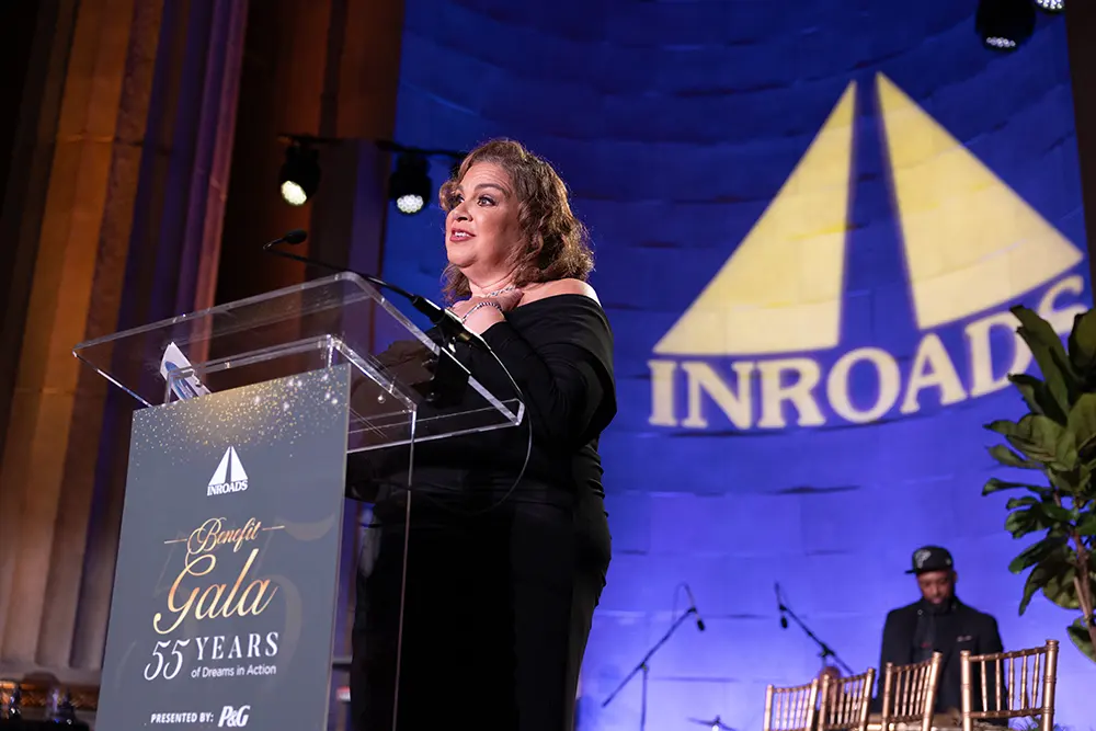 A woman in a black dress stands at a podium with the INROADS logo and “Annual Gala 55 Years” written on it. A large screen is behind her with a deep purple background displaying the INROADS name and logo in a cream color.