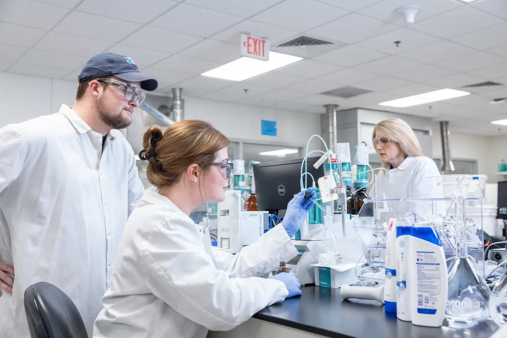 Three scientists in lab coats and safety glasses are working in a lab, with one woman pipetting.