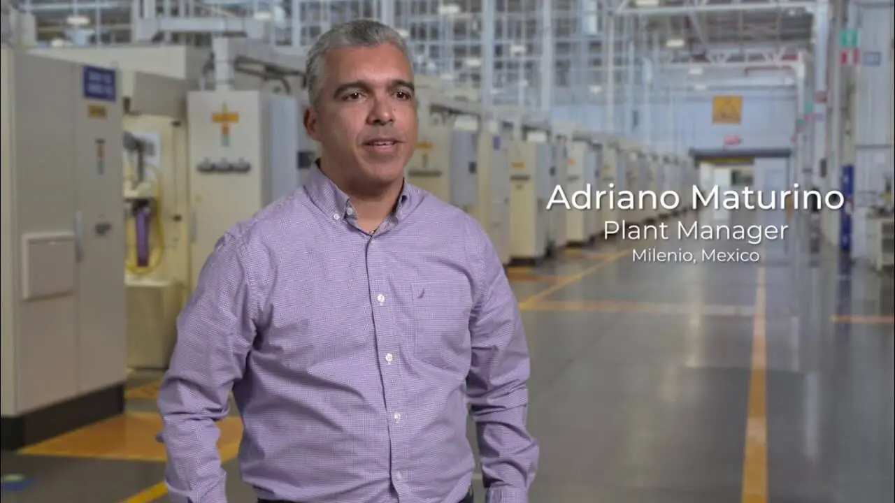 A man in a long sleeve lilac shirt. He stands inside a large manufacturing plant.