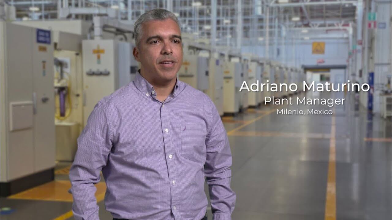 A man in a long sleeve lilac shirt. He stands inside a large manufacturing plant.