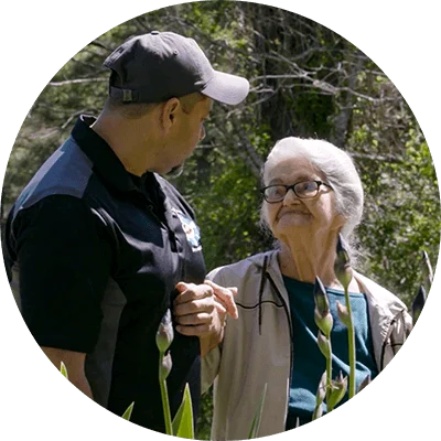 A man and an elderly woman hold hands while they stand outside in front of a patch of long stemmed purple flowers. Numerous trees are lined up across the background.