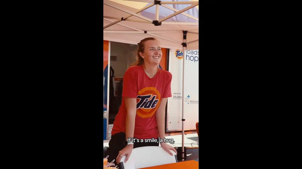 Smiling woman wearing a red Tide T-shirt stands behind a table under a canopy at a community outreach event