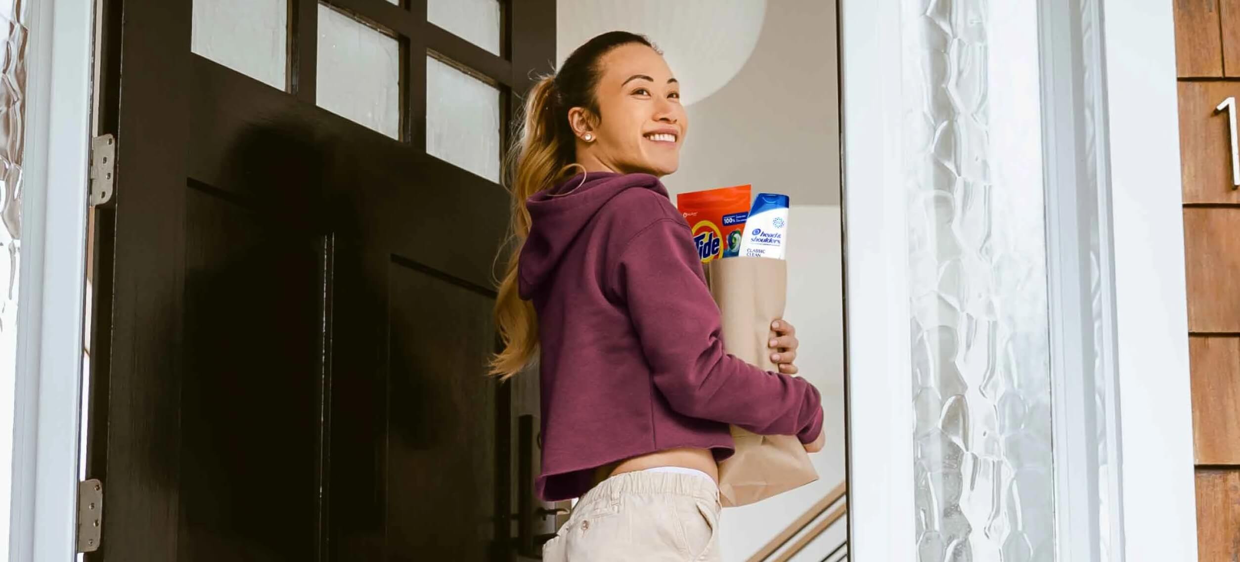 Woman enters front door of house while holding brown paper grocery bag containing Tide detergent and Head & Shoulders shampoo 