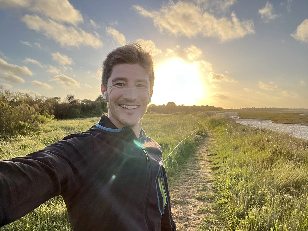A smiling man in a dark athletic jacket stands in a grassy field, with a sunset sky behind him.