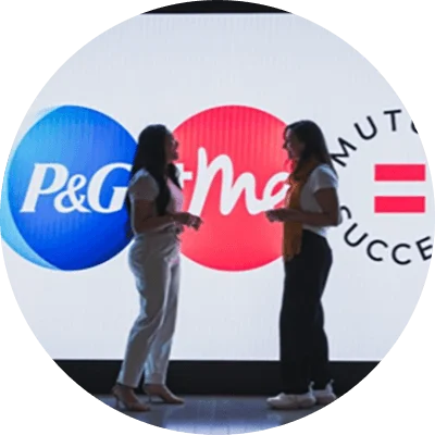 Two women face each other as they engage in conversation. They are standing at the end of a hallway, in front of a digital display of a blue, white and pink logo.