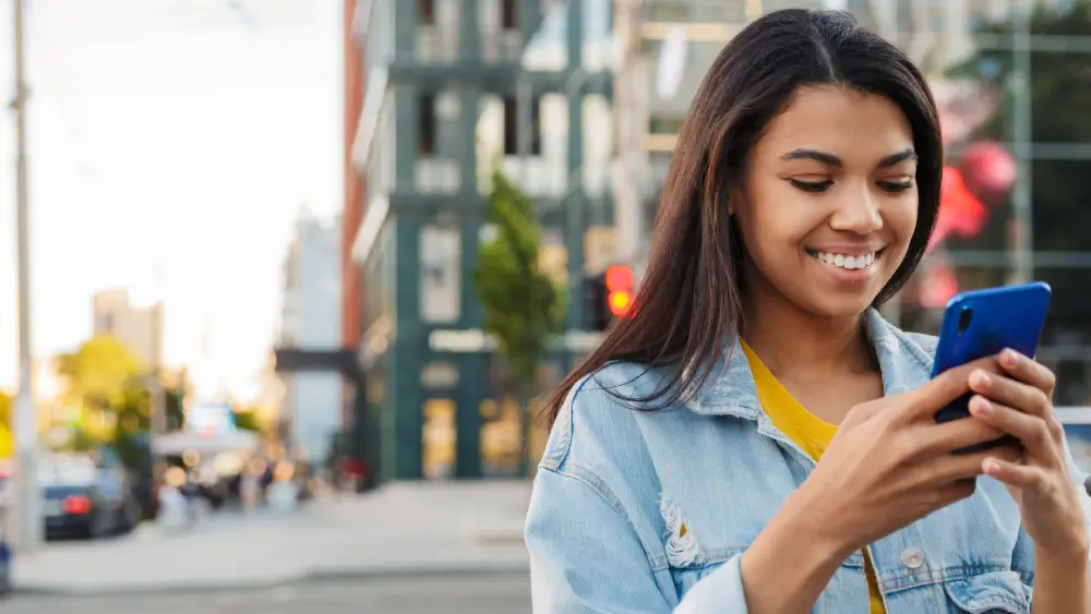 A smiling young woman in a denim jacket looks at her blue smartphone on a blurred city street.