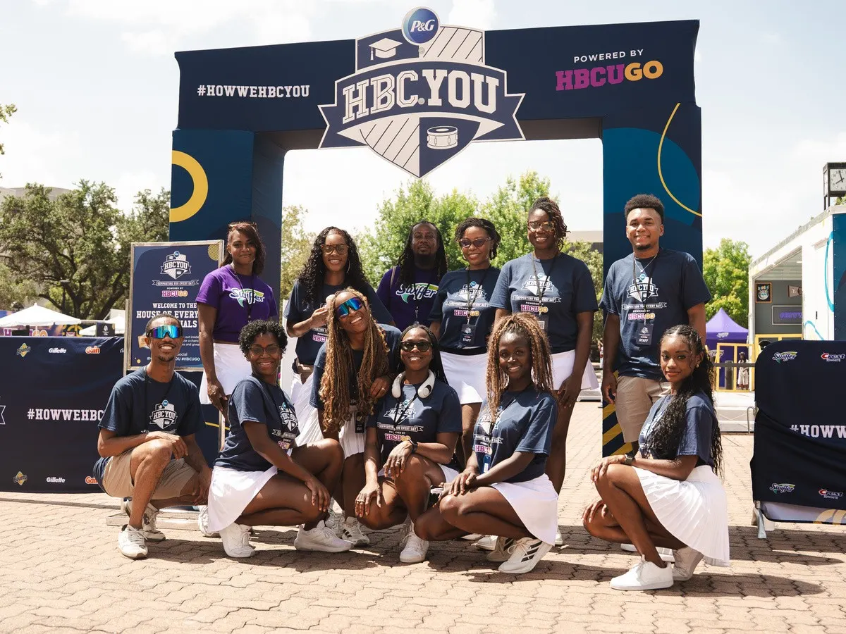 Eleven students in blue shirts stand together outdoors in front of a large branded display.