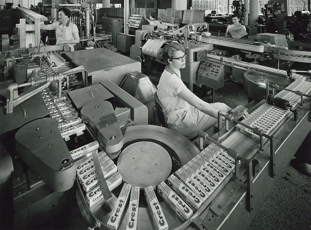Archival image of a factory where women are packaging Crest toothpaste. Conveyors transport the boxes