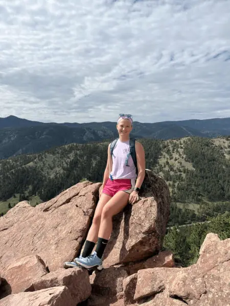 A young white woman sitting atop a large rock with mountains in the background.