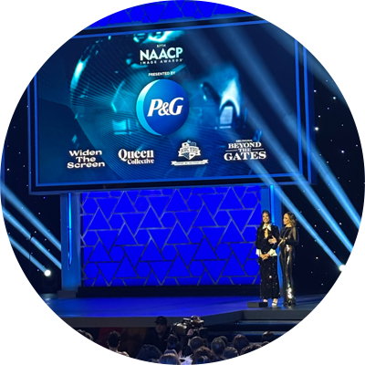 Two cast members from Beyond the Gates in black dresses stand on stage at the 57th NAACP Image Awards. A large blue screen behind displays P&G’s logo as sponsors and Widen The Screen, Queen Collective, HBCYOU, and Beyond the Gates word marks/logos. 
