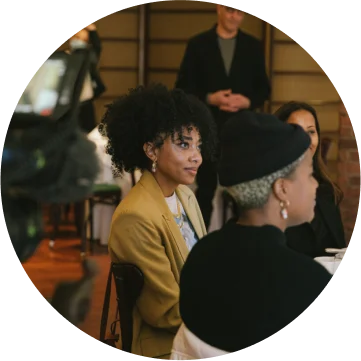 Three women are sitting at a table and are looking towards something in the front of the room. 