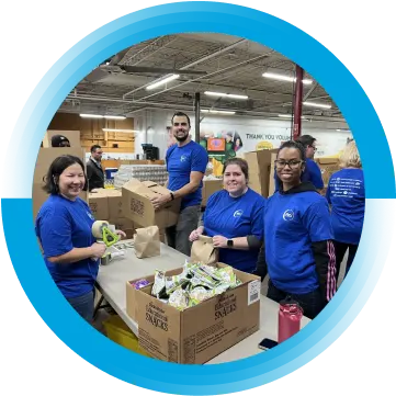 Four people wearing dark blue t-shirts with the P&G logo on them are packing snacks into paper bags.