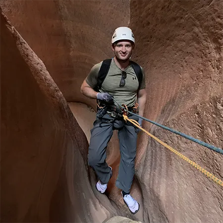 An adventurer in outdoor gear and a white helmet descends into a narrow, orange-walled canyon using ropes and a harness.