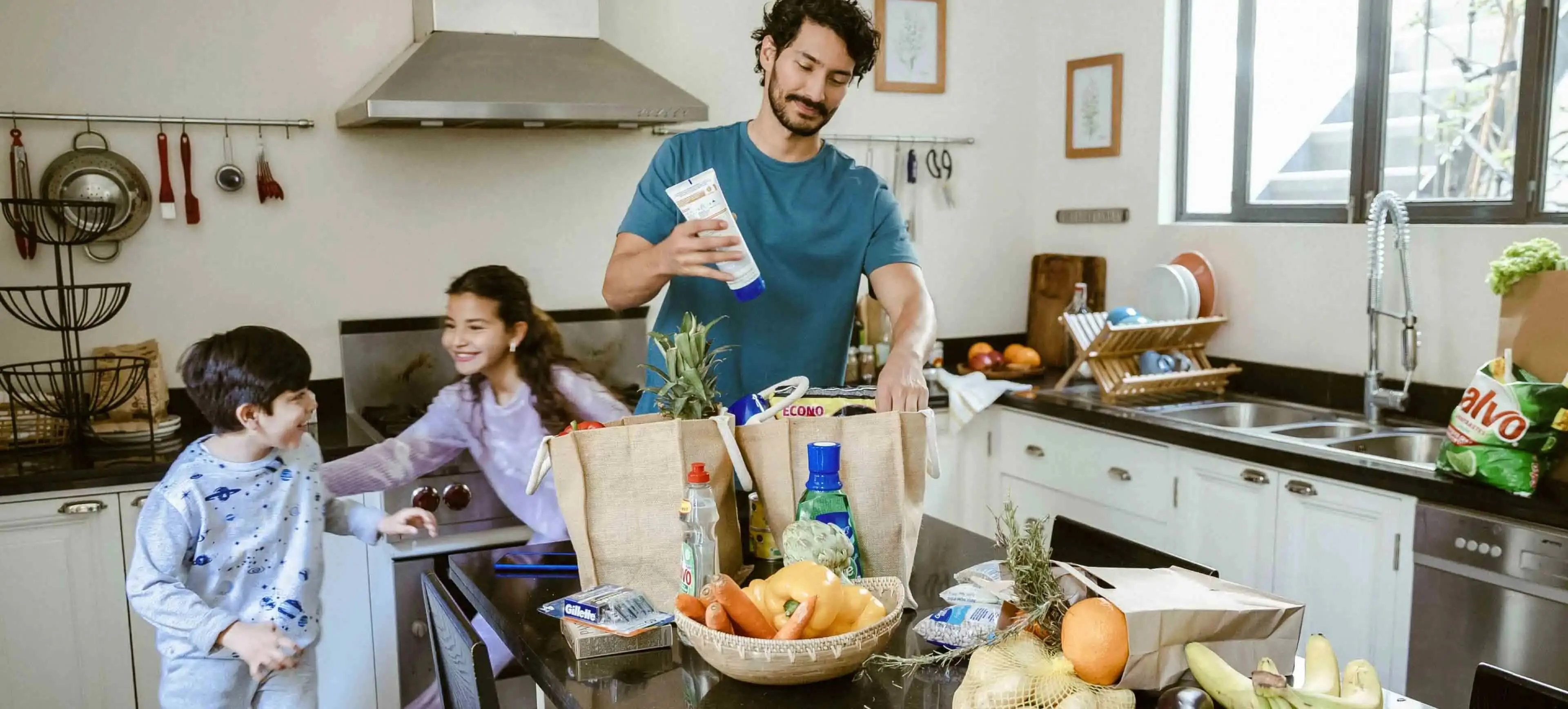 Man is unpacking groceries in kitchen while two children play in the background
