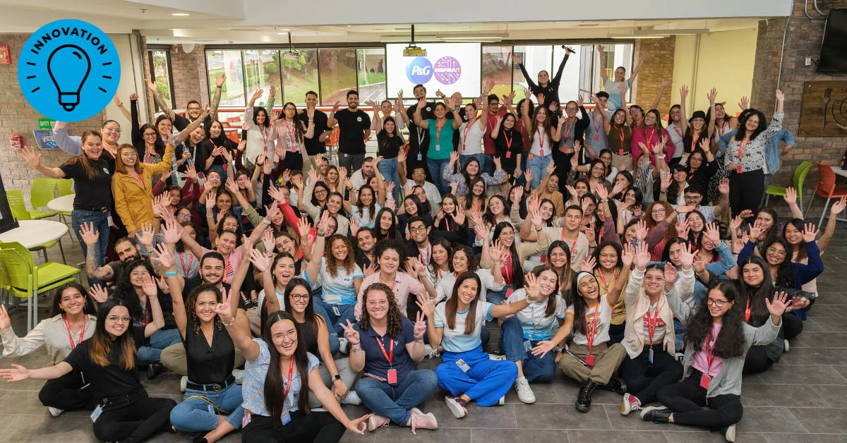 A large group of Hispanic Procter & Gamble employees pose, with many sitting on the floor and others standing in the background. They are waving and smiling. The group is predominantly female.
