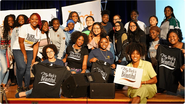 A group of twenty two women are posing for a group photo, holding up a sign that say “My Black is Beautiful.” 