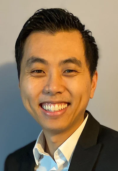 An Asian man with short black hair is wearing a white button up shirt and a black jacket as he smiles directly at the camera.