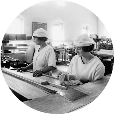 Two young women in white caps and uniforms. They stand in a manufacturing facility behind a long table with a long row of soap bars. The photo is from 1910.