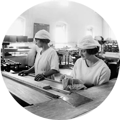 Two young women in white caps and uniforms. They stand in a manufacturing facility behind a long table with a long row of soap bars. The photo is from 1910.