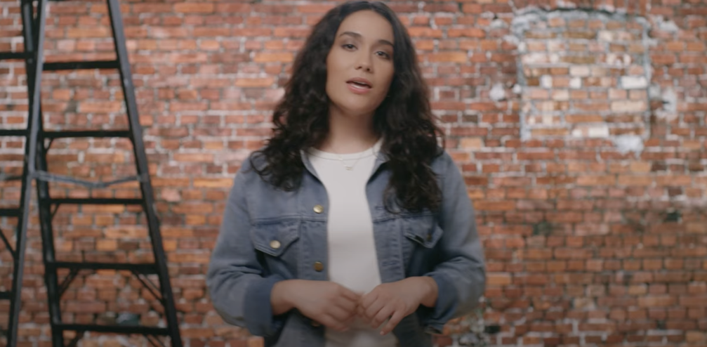 Woman with long brown hair talking and looking towards the camera. She is wearing a white top and a denim jacket. She is standing in front of a brick wall.