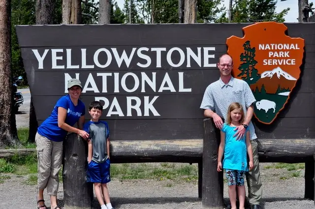 An adult female, an adult male and a young boy and girl stand together in front of a large wood sign that says "Yellowstone national park. National park service."