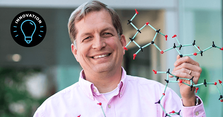 A man with short blonde hair wears a pink buttoned shirt. He smiles as he holds up a plastic molecular model.