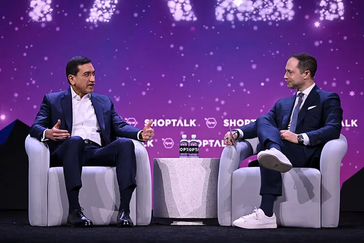 Two men in dark blue business suits sit together on a stage while having a discussion. A purple digital background also displays a white text logo that reads, "Shoptalk."