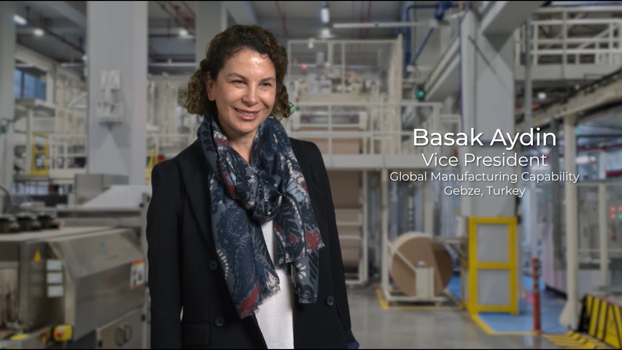 A woman with short curly haired. She wears a dark jacket and scarf and stands inside a manufacturing plant.