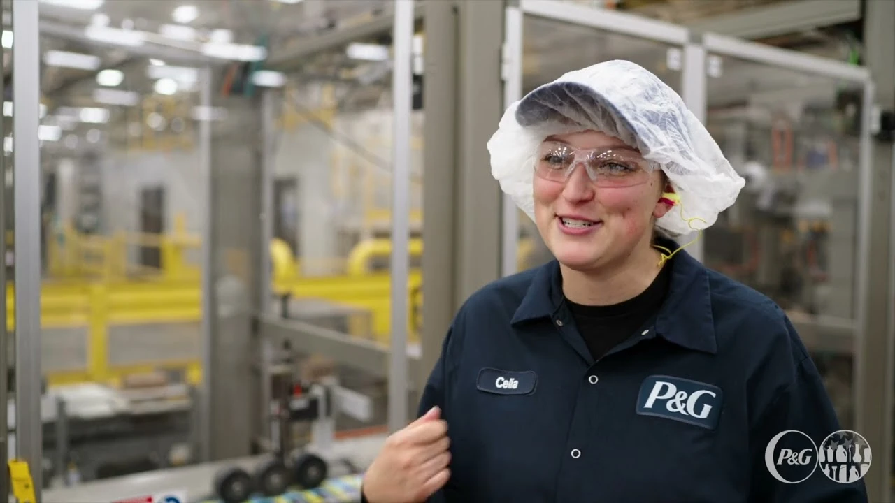 Factory worker wearing safety glasses, hair covering, and ear protection smiling in an industrial manufacturing facility while wearing a P&G uniform.