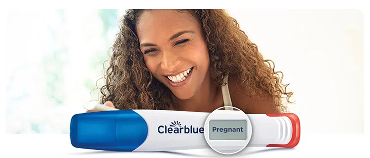 A woman with tan skin and long curly brown hair, stands against a white background. In the foreground, there is a white and blue Clearblue pregnancy test with the digital display showing the word 'pregnant.' The woman's expression suggests joy.
